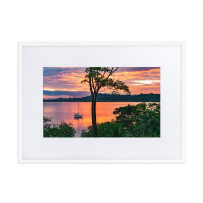 Sailboat anchored in a calm, jungle-fringed bay with an orange and red sky.