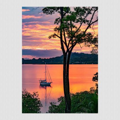 Sailboat anchored in a calm, jungle-fringed bay with an orange and red sky.