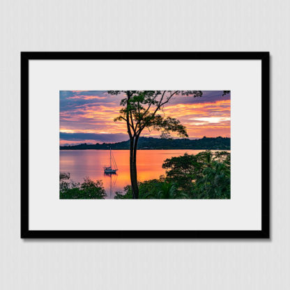 Sailboat anchored in a calm, jungle-fringed bay with an orange and red sky.